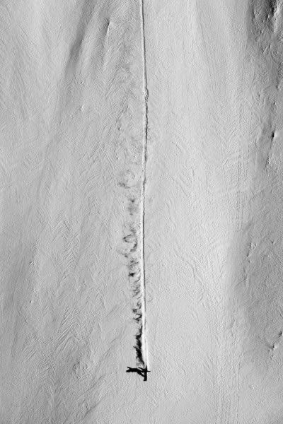 An aerial black and white photograph of a wide snow face, with a single ski track running the full height of the frame and the skier a small dark figure at the end of the line.