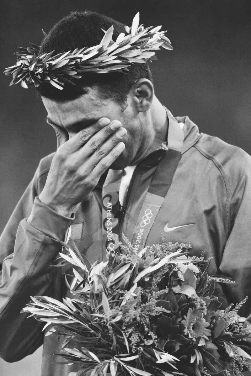 An Olympic medallist on the podium in Athens wearing an olive wreath, one hand covering his face, photographed in black and white.